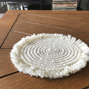 Detailed close-up of a cream-white braided cotton coaster with fringes on a wooden table