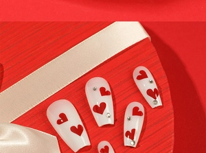 Detailed view of white trapezoid nails with red hearts and rhinestones on a red gift box