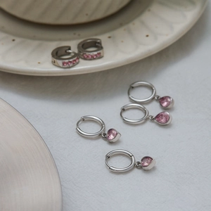 Detail of silver hoop earrings with pink zircons and heart pendants on white fabric