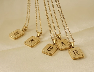 Primer plano de collares de letras en acero de titanio dorado con dijes cuadrados mate