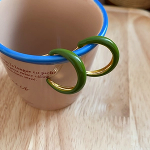 Close-up of dark green enamel C-shaped hoop earrings hanging on a pink ceramic cup
