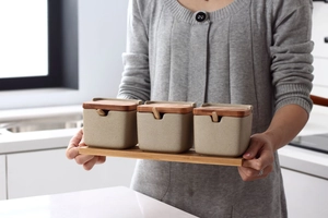 Ceramic seasoning jar set with wooden lids and spoons on a tray in kitchen hands