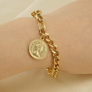 Bracciale a catena dorata con ciondolo a moneta Elizabeth indossato su sfondo di raso caldo