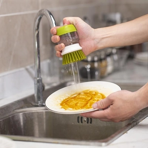 Green handheld dish brush with soap dispenser scrubbing stains on a ceramic plate