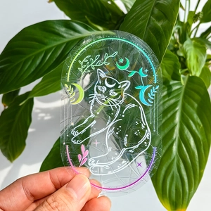 Holographic suncatcher sticker with cat moon and star line art held against green plants