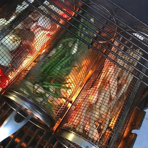 Cylindrical stainless steel BBQ rolling grill basket heating vegetables over hot charcoal