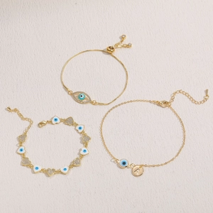 Three gold evil eye bracelets with zircon inlay and heart charms on a beige texture surface