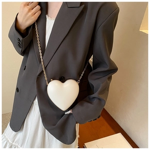 White heart-shaped crossbody bag with gold chain strap worn with a grey blazer and dress