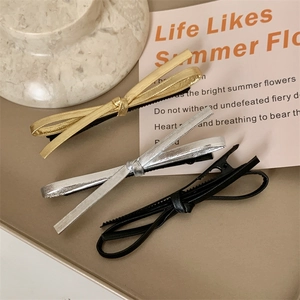 Three minimalist leather bow hair clips in gold, silver, and black on a beige display tray