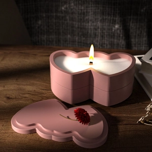 Pink heart-shaped candle cup with burning wood wick and floral lid on wooden desk