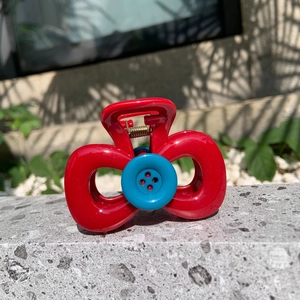 Elegant red bow-shaped hair claw clip with a blue button accent on a stone surface