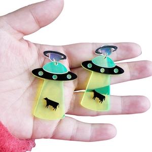 Detailed close-up of acrylic UFO earrings featuring a green beam lifting a small black cow