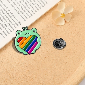 Cute green cartoon frog enamel pin holding a rainbow heart on an orange surface