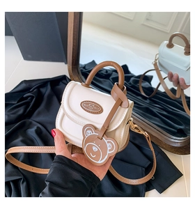 Detail of a mini cream saddle bag with brown handles and a cute bear charm accessory