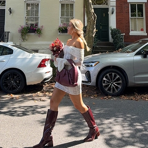 Street style shot of a white lace off-shoulder mini dress with burgundy boots and a handbag