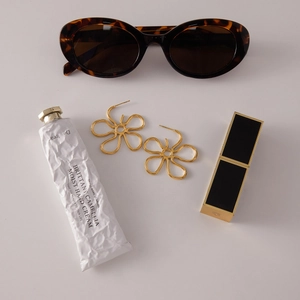 Flat lay of gold flower earrings paired with tortoiseshell sunglasses and cosmetics