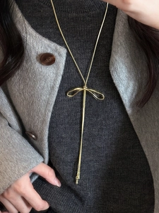 Detail showing a golden bow necklace with long snake chain tassels on a gray sweater