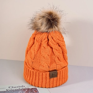 Close-up of orange cable knit beanie featuring a dark pompom and a leather logo patch