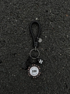 Detail of woven leather keychain with black poker chip, dice, and heart charm on asphalt