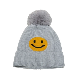 Close-up of grey knit beanie featuring a yellow smiley face and large faux fur pom-pom