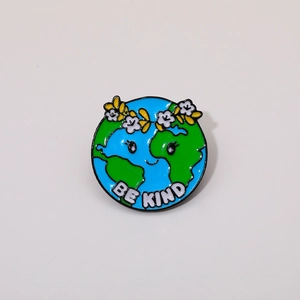 Detail of a cartoon enamel pin featuring a smiling Earth and Be Kind slogan