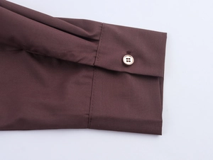 Detail shot of a dark brown long-sleeve cuff with a gold-edged button on cotton fabric