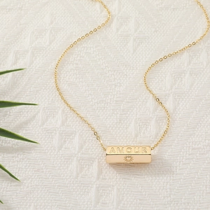 Golden necklace with AMOUR engraved rectangular pendant on white textured fabric background