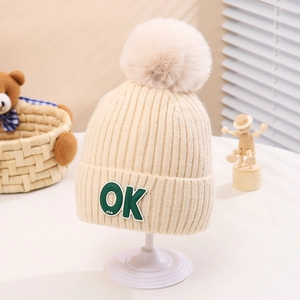 Close-up of knit texture on a beige kids beanie with OK patch and large pom-pom detail