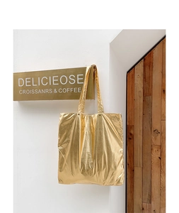 Detailed close-up of a large golden metallic tote bag hanging on a minimalist white wall