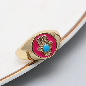 Close-up of golden Hamsa ring with pink enamel and blue evil eye detail on white ceramic