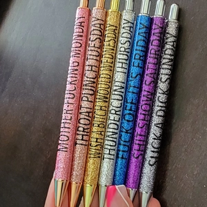 Detail showing 7 glitter ballpoint pens with bold text labels and colorful finishes