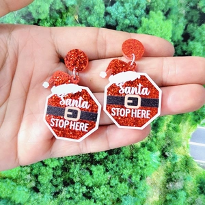 Detail of red glitter acrylic Santa Stop Here earrings held against greenery