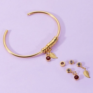 Detail of gold open bangle with red gemstone charm and gold leaf pendant on purple background