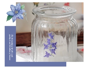 Floral print glass candle jar with purple blooms on a soft focus wooden desk scene