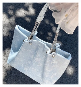 Detail of light blue denim tote bag featuring a silver metal chain strap on pavement