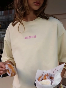 Dettaglio della maglietta in cotone crema con stampa ASSERTIVE rosa e spalle scese