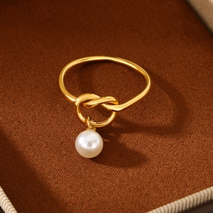 Detail of a minimalist gold ring with a knot design and a hanging white pearl in a box