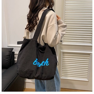 Detail of a black canvas tote bag with blue Beach text worn by a model in a minimal room