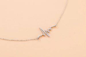 Macro detail of silver necklace featuring heartbeat ECG line pendant on pink background