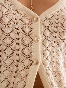 Gros plan détaillé sur la maille ajourée et les boutons coquillages dorés du gilet blanc