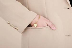 Détail d'un bracelet doré avec breloque cœur gravée MOM porté au poignet avec un costume beige