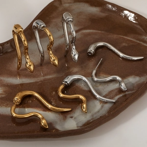Minimalist snake-shaped stainless steel earrings in gold and silver finish set on a ceramic tray