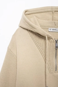 Detail of beige knitted hoodie neckline with silver zipper, drawstring, and knit texture