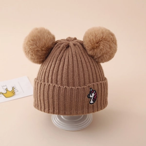 Detail of a brown ribbed knit hat with dual pom poms and a colorful unicorn embroidery