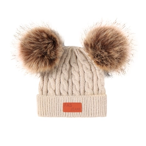 Close-up of beige knit baby hat with twin brown faux fur pom-poms and orange leather tag