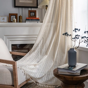 Close-up of beige knit lace curtain with fringe tassels in a cozy bedroom setting