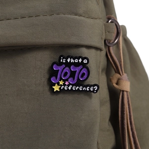 JOJO enamel pin on a green canvas bag featuring purple letters and yellow stars