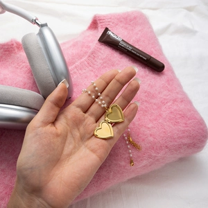 Détail d'un médaillon cœur doré ouvert avec chaîne en perles tenu sur un pull rose doux