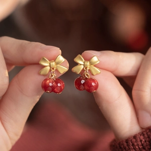 Detail view of matte gold bow earrings with dark red cherry beads held by hand