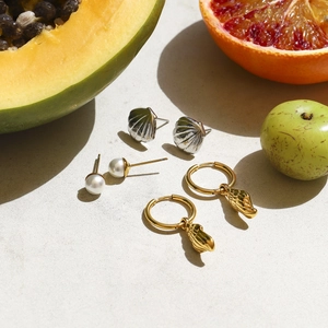 Flat lay of gold shell earrings and pearl studs on stone background with fresh fruit